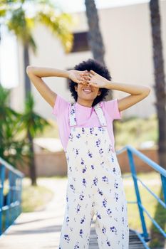 Carefree female in casual clothes covering eye with hands while standing on walkway against buildings on blurred background on summer day