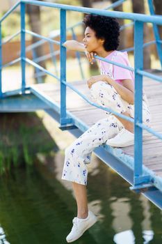 Full body side view of focused African American female sitting on wooden footbridge over lake on summer day in park