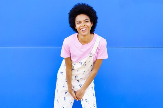 Optimistic African American female with lack hair in casual clothes leaning forward and looking at camera against blue background on street