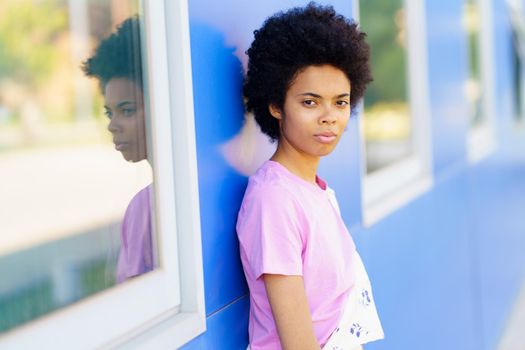 Serious African American female with Afro hairstyle looking away with thoughtful face while standing near blue building with windows on street of city