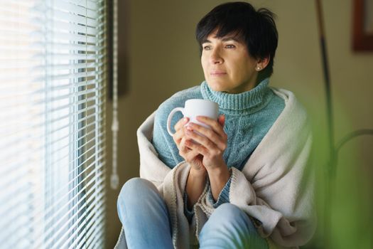 Mid adult woman drinking coffee and looking out of the window in winter day. Female in her 50s