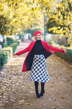 Full body of cheerful female in red coat and beret looking at camera while spinning around on walkway in autumn park