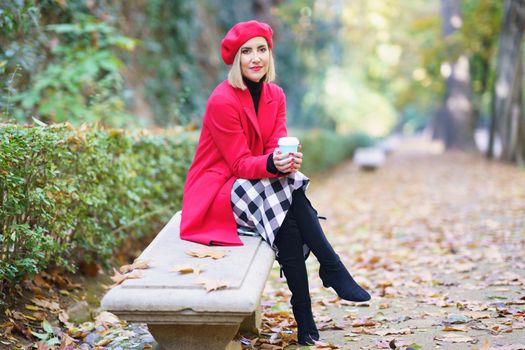 Full body of glad female in elegant beret and coat, with paper cup of takeaway hot drink, looking at camera while sitting on bench in park