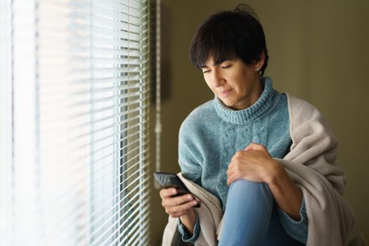 Middle-aged woman consulting something on her smartphone near the window at home. Female in her 50s