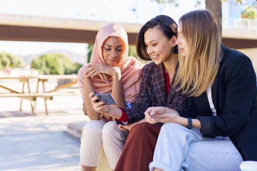 Content young multiracial female best friends, in casual clothes and hijab, smiling while watching video on smartphone sitting on bench in city park on sunny day