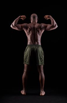 Rear view of an African American muscular man flexing arm muscles on a black background. Athlete adult man standing with shorts in a studio.