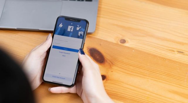 CHIANG MAI ,THAILAND - AUG 18, 2022 : Woman hand holding iPhone X to use facebook with new login screen.Facebook is a largest social network and most popular social networking site in the world..