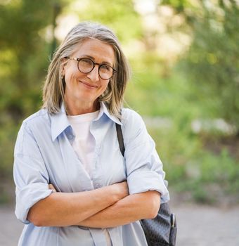 Beautiful grey hair mature woman wearing glasses and backpack standing arms folded outdoor enjoying vacation traveling around world at retirement. Mature woman with perfect skin wearing blue shirt.