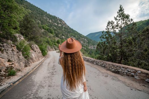 Female traveler in brown hat and white dress looking at amazing mountains and forest, wanderlust travel concept, atmospheric epic moment.
