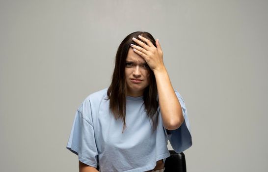 Portrait of stressed beautiful brunette woman in a blue t-shirt holding her head with hands having headache over grey wall. Attractive female suffering from head pain