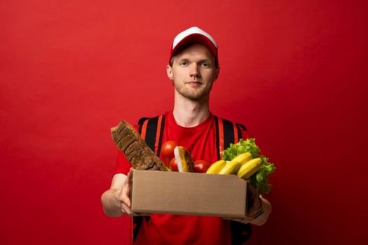 Delivery employee in red t-shirt and cap working courier service from shop restaurant to home holding brown craft paper takeaway food box with vegetables