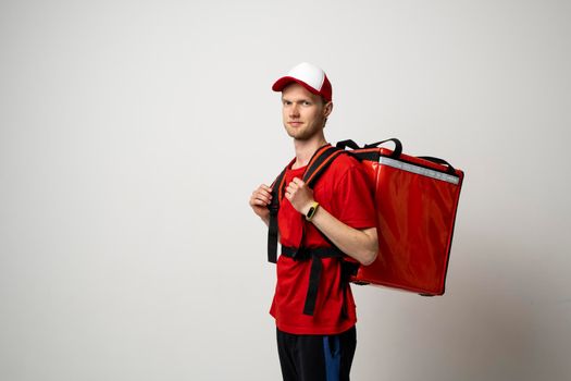 Delivery man in a red uniform and a cap carry red thermal backpack full of food, groceries to the clients on a white background. Food delivery service