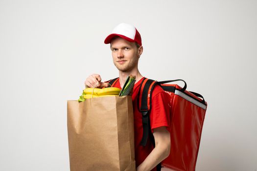 Smiling delivery employee man 20s in red t-shirt working courier service from shop restaurant to home holding brown craft paper takeaway food bag mockup
