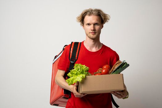 Delivery employee in red t-shirt working courier service from shop restaurant to home holding brown craft paper takeaway food box with vegetables
