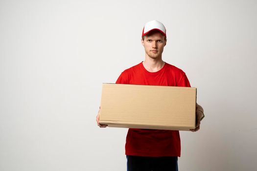 Young delivery man, courier in a red uniform with a big parcel in a hand over white background