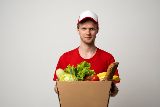Smiling delivery employee in red t-shirt working courier service from shop restaurant to home holding brown craft paper takeaway food box mockup