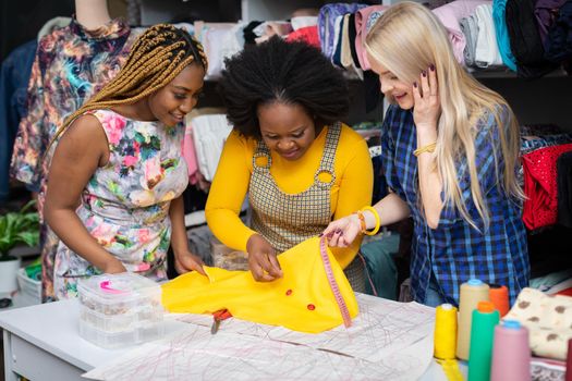 Creative dark-skinned girls and one European woman match buttons to a selected sewing fabric. Two African girls and a European woman in a sewing studio.
