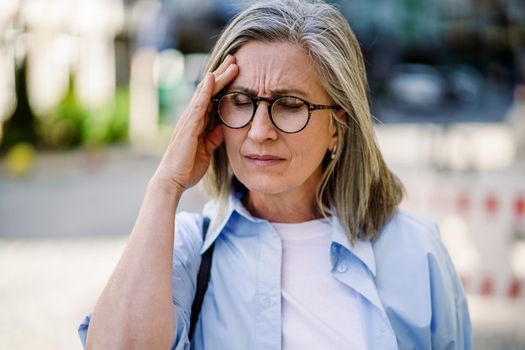 Feeling hard headache, migraine mature grey haired woman 50s, 60s, standing outdoors holding her head. Suffering from summer teat feeling sick while travel mature woman standing outdoor.