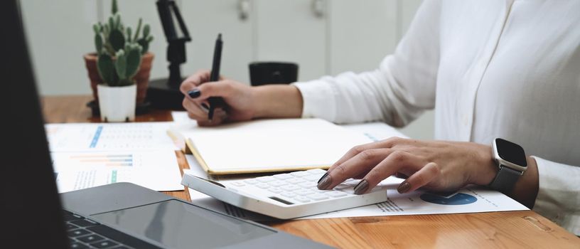 accountant working on calculator and laptop computer to calculate business data during make note at notepad, accountancy document at office.