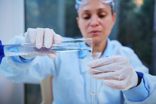 Details: Graduated cylinder in the gloved hands of a blurred woman scientist, lab technician pouring transparent liquid into a test tube through a laboratory funnel in a medical science laboratory
