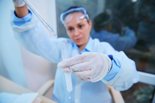 Selective focus on a test tube with a taking place chemical reaction and graduated lab pipette in a blurred woman scientist hands dripping reagent, conducting science experiment in research laboratory