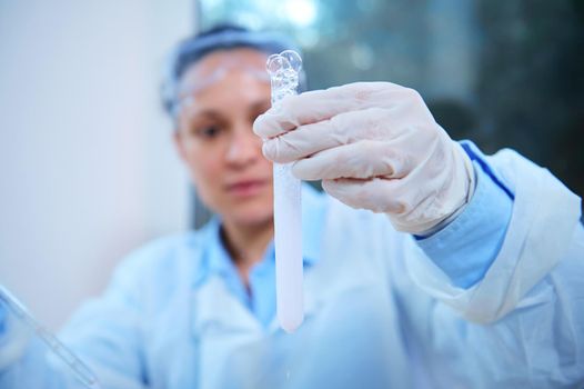 Details: A test tube in the hands in protective gloves of a blurred background of a scientist pharmacologist examining the taking place chemical reaction in a science clinical research laboratory