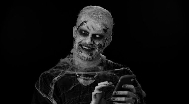 Scary man with Halloween zombie bloody wounded makeup using mobile phone typing new post on web, sms message, browsing, addiction of social networks. Sinister one undead guy on black wall background