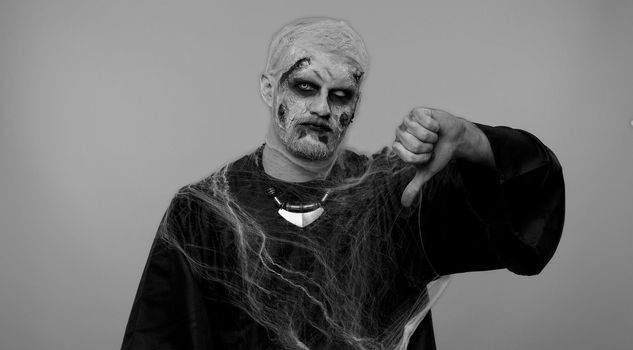 Sinister man in carnival costume of Halloween crazy zombie with bloody wounded scars face showing thumbs down sign gesture, expressing discontent, disapproval, dissatisfied, dislike. Cosplay undead