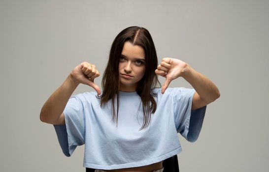 Young brunette woman in a oversized blue t-shirt isolated on grey background showing thumb down gesture with two hands