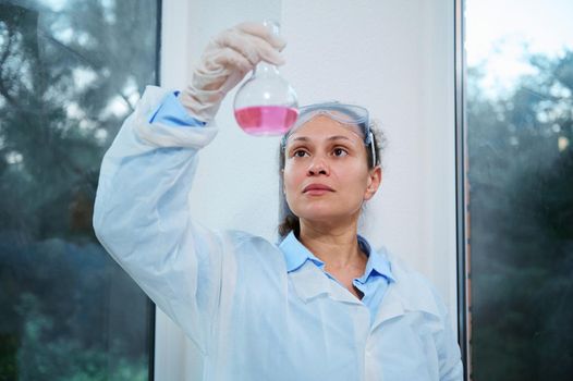 Beautiful multi-ethnic woman scientist, medical biologist, pharmacologist in lab coat examines the fluid substance in glass flask while making clinical study in medical scientific research laboratory