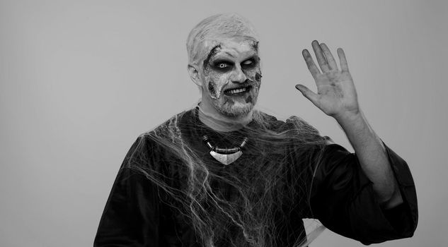Creepy man with bloody scars face, Halloween zombie make-up. Scary wounded undead guy smiling friendly at camera and waving hands gesturing hello or goodbye, welcoming with hospitable expression