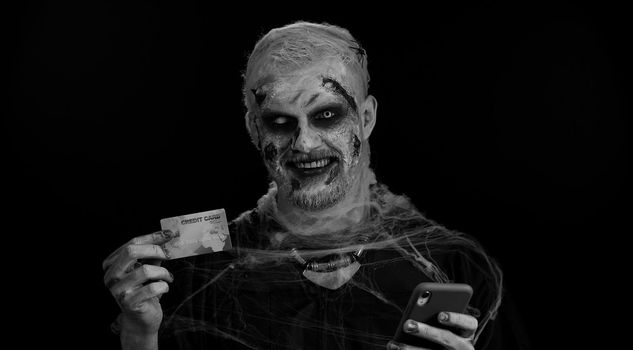 Sinister man with horrible scary Halloween zombie make-up using credit bank card and smartphone while transferring money, purchases online shopping. Dead guy with wounded bloody scars face, black room