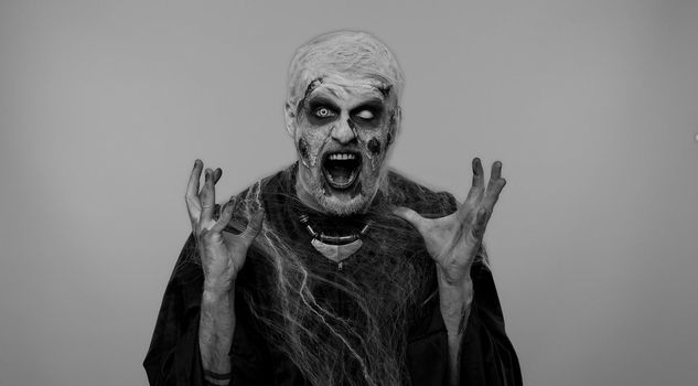 Scary man in carnival costume of Halloween crazy zombie with bloody wounded scars face screaming, shouting, trying to scare on gray. Horror theme of cosplay wounded undead, beast, monster