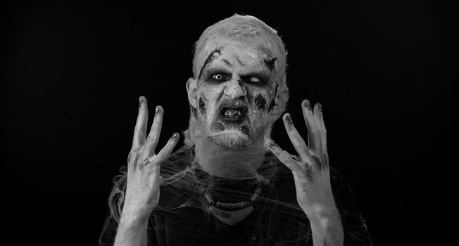 Zombie man in convulsions with makeup with fake wounds scars and white contact lenses trying to scare in dark studio room. Sinister dead guy hiding through hands. Halloween, filming, staging concept