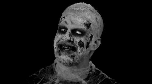 Smiling man with horrible scary Halloween zombie makeup in convulsions making faces, looking ominous, trying to scare. Dead guy with wounded bloody scars face isolated against black wall background