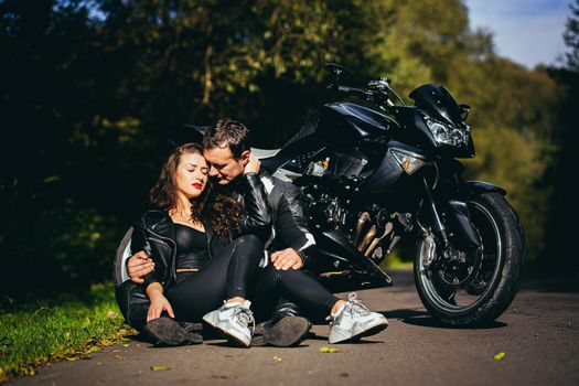 Handsome young man and beautiful young girl, couple sitting near motorcycle in black leather clothes, hugging, in nature, outdoors