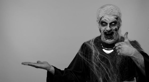 Creepy man with bloody scars face, Halloween stylish zombie make-up. Scary wounded undead guy showing thumbs up and pointing at right on blank space. Place for advertisement logo. Sinister monster