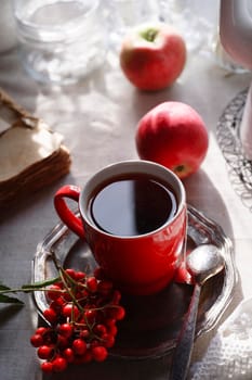 Red cup of tea on vintage metal plate with a bunch of red rowan and two red apples, autumn morning tea concept, selective focus.