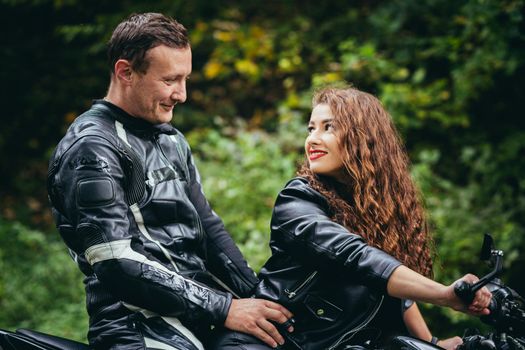 Handsome young man and beautiful young girl, couple sitting on a motorcycle in black leather clothes, close up, hugging, in nature, outdoors, looking at camera