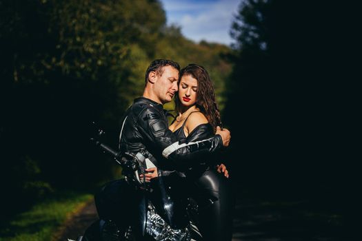 Handsome young man and beautiful young girl, couple sitting on a motorcycle, standing near a motorcycle in black leather clothes, hugging, in nature, on the street