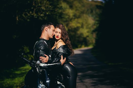 Handsome young man and beautiful young girl, couple sitting on a motorcycle, standing near a motorcycle in black leather clothes, hugging, in nature, on the street