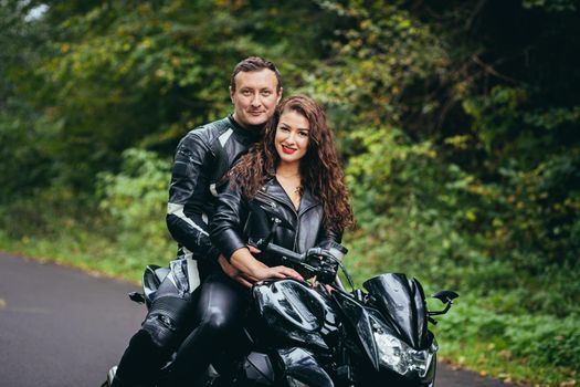 Handsome young man and beautiful young girl, couple sitting on a motorcycle in black leather clothes, close up, hugging, in nature, outdoors, looking at camera