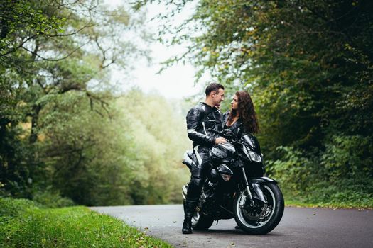 Handsome young man and beautiful young girl, couple sitting on a motorcycle, standing near a motorcycle in black leather clothes, hugging, in nature, on the street