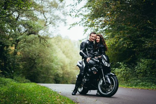 Handsome young man and beautiful young girl, couple sitting on a motorcycle, standing near a motorcycle in black leather clothes, hugging, in nature, on the street