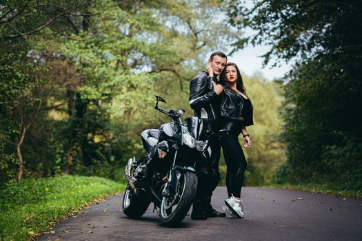 Handsome young man and beautiful young girl, couple sitting on a motorcycle, standing near a motorcycle in black leather clothes, hugging, in nature, on the street