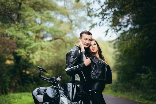 Handsome young man and beautiful young girl, couple sitting on a motorcycle, standing near a motorcycle in black leather clothes, hugging, in nature, on the street