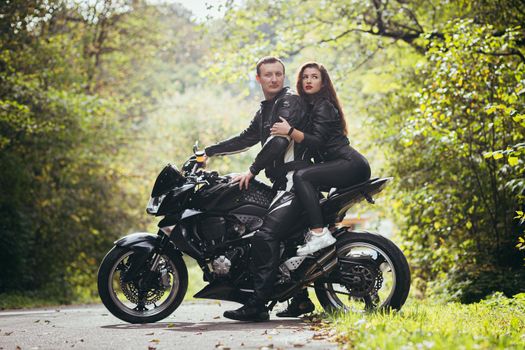 Handsome young man and beautiful young girl, couple sitting on a motorcycle, standing near a motorcycle in black leather clothes, hugging, in nature, on the street