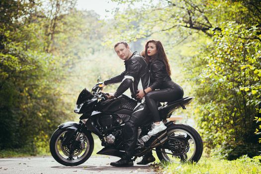 Handsome young man and beautiful young girl, couple sitting on a motorcycle, standing near a motorcycle in black leather clothes, hugging, in nature, on the street