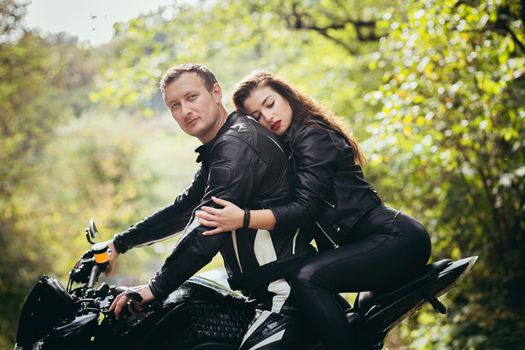 Handsome young man and beautiful young girl, couple sitting on a motorcycle in black leather clothes, close up, hugging, in nature, outdoors, looking at camera