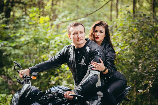 Handsome young man and beautiful young girl, couple sitting on a motorcycle in black leather clothes, close up, hugging, in nature, outdoors, looking at camera